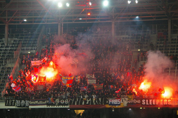 Eine große Menge in einem Stadion hält Fahnen und Banner, mit Leuchtraketen, die Rauch erzeugen, unter einer Decke mit Deckenleuchten und Metallrahmen.