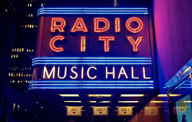 Außensicht des Radio City Music Hall in New York City bei Nacht, hell erleuchtet mit hohen Gebäuden im Hintergrund, mit einer 'Radio City'-Leuchtschrift und zusätzlichen Schildern auf der rechten Seite.
