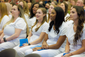 Eine Gruppe von Frauen in weißen Kleidern sitzt in einem Hörsaal, jede hält ein blaues Objekt, mit einem unscharfen Hintergrund, der die Frauen hervorhebt, die scheinbar Teil eines Pflegeprogramms sind.