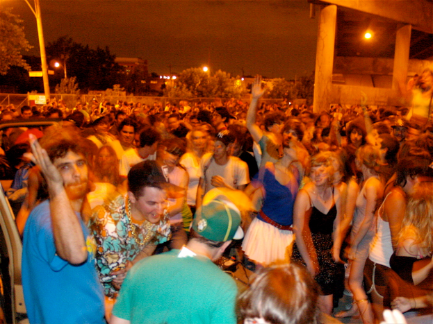Menschen tanzen im Vordergrund auf einem Konzert, mit Lichtern, Polen, Bäumen und anderen Gegenständen im Hintergrund.