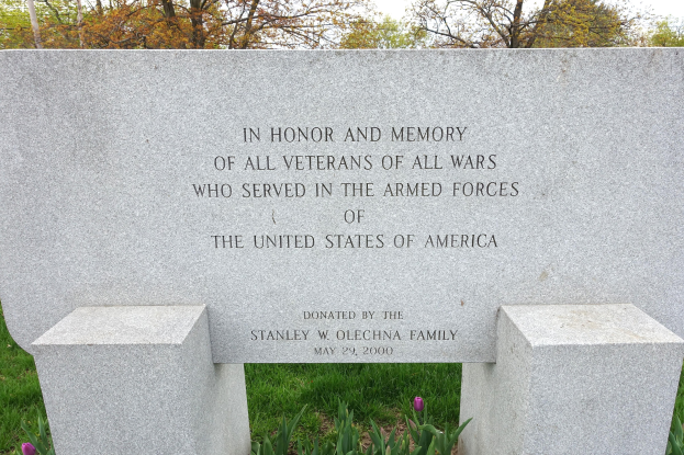 Gedenkstein mit Inschrift für US-Streitkräfte-Veteranen, umgeben von Pflanzen, Blumen, Bäumen und einer bewölkten Himmel