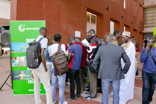 Eine Gruppe von Menschen steht auf einem Gehweg, wobei eine Person in einem blauen T-Shirt vorbeigeht, eine andere ein Buch in schwarzer Kleidung hält und zwei mit Rucksäcken; ein Banner mit Text, Bildern und einem Gebäudeabbild ist sichtbar, sowie ein Lautsprecher, Metallstangen, ein Fahrzeug und Gebäude im Hintergrund.