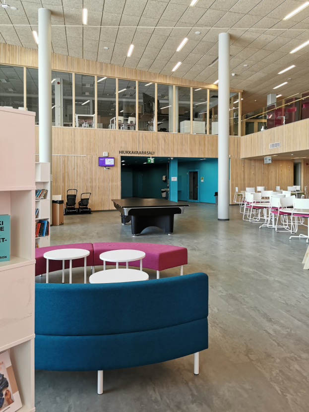 Großer Raum mit mehreren Tischen, Stühlen, Sofas, Hockern, Bücherregalen, Deckenlampen, einem wandmontierten Fernseher und Säulen, der an das Innere einer Schulbibliothek erinnert.