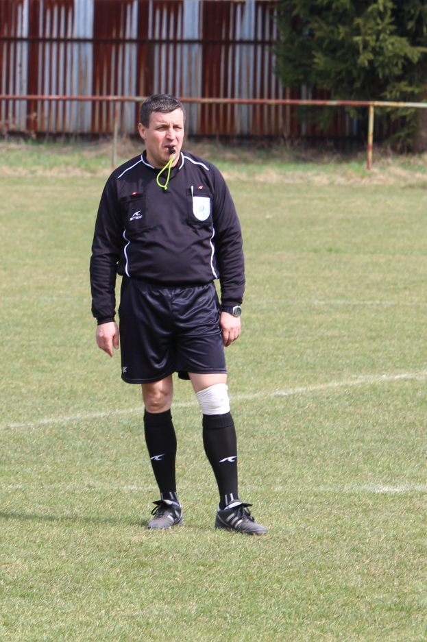 Ein Fußball-Schiedsrichter in Uniform steht auf einem Feld umgeben von einem Zaun und Bäumen.