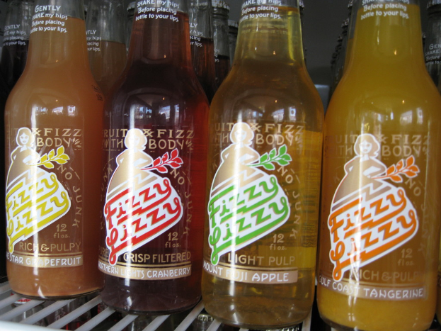 Drei farbenfrohe Getänkeflaschen mit der Aufschrift 'Fizzy Lizzy' in Gelb, Rot und Orange auf einem Tisch.