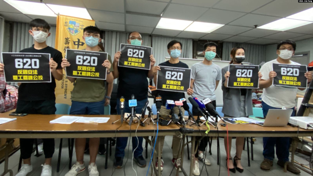 Eine Gruppe maskierter Personen mit Schildern steht vor einem Tisch mit Mikrofonen, Papieren und Laptops und protestiert gegen das Auslieferungsgesetz in Hong Kong, mit einem Banner und einer beleuchteten Decke im Hintergrund.