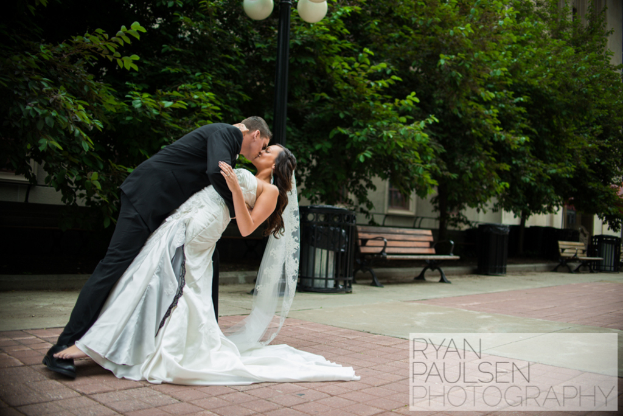 Eine Braut und ein Bräutigam küssen sich vor Bänken, Mülltonnen und Bäumen.