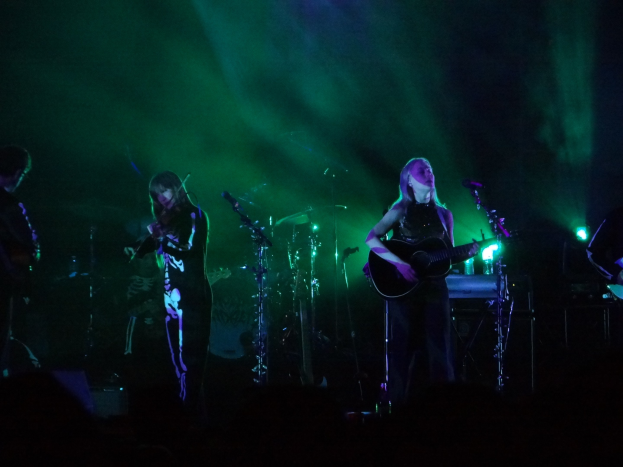 Eine Gruppe von Menschen auf der Bühne mit Gitarren, Mikrofonen und Musikinstrumenten in einer schwach beleuchteten Umgebung.