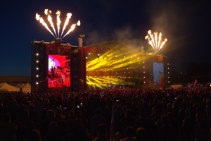 Eine große Menschenmenge auf einem Musikfestival mit einer hell erleuchteten Bühne mit Performern und Bannern, Zelte links und Feuerwerk am dunklen Himmel.
