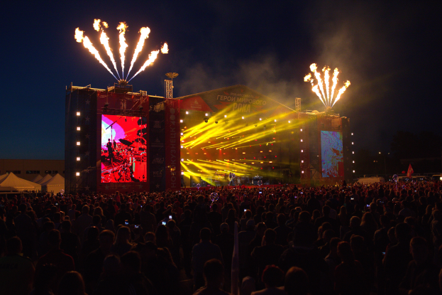 Eine große Menschenmenge auf einem Musikfestival mit einer hell erleuchteten Bühne mit Performern und Bannern, Zelte links und Feuerwerk am dunklen Himmel.