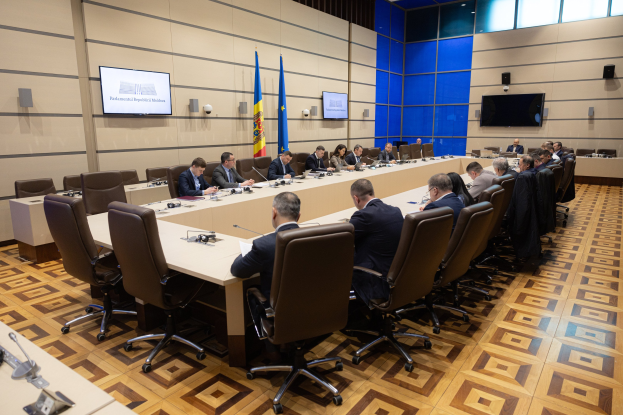 Große Gruppe von Menschen um einen langen Tisch mit Mikrofonen und Papieren in einem offiziellen Konferenzraum mit Flaggen der Europäischen Union, Fernsehern und Deckenleuchten.