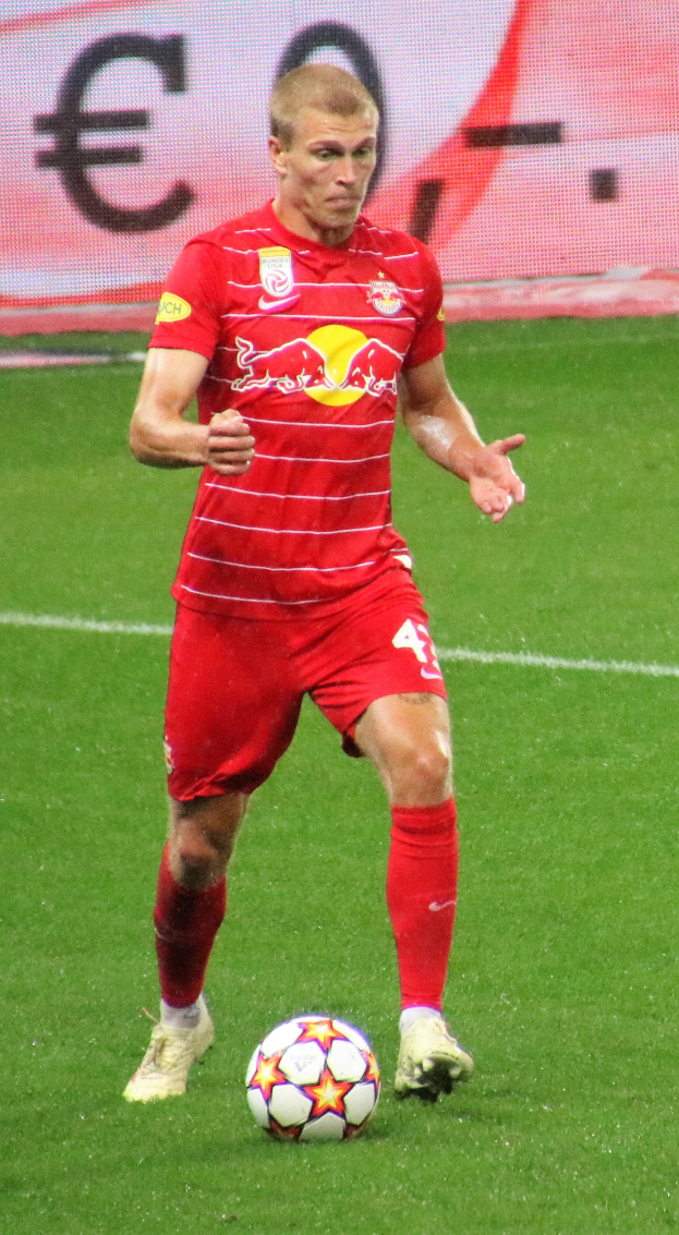 Mann in rotem Trikot spielt Fussball auf einem Feld, mit einer Tafel im Hintergrund, trägt Fussballausrüstung und -schuhe.