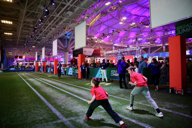 Zwei junge Jungs spielen Fußball auf einem Feld, umgeben von einer Gruppe von Menschen, mit Flaschen und anderen Gegenständen auf der rechten Seite, einem Zaun, Bannern, einem Anzeigebildschirm, Metallrahmen, einem Dach mit Deckenleuchten und Metallpfosten im Hintergrund.