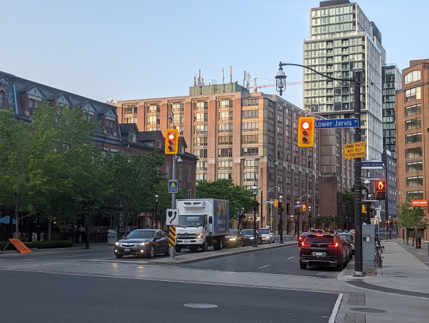 Eine stark befahrene Stadtstraße mit einem Lastwagen, einem Auto und einem SUV, umgeben von Laternen, Ampeln, Anzeigetafeln, Bäumen und Gebäuden unter einem Himmel.