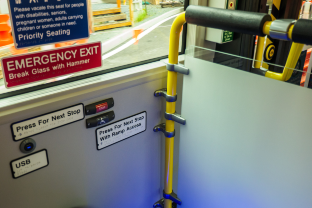 Notausgangsschild an der Seite eines Zuges, mit einer Tür mit Griff im Vordergrund und einem Glasfenster im Hintergrund, das Straße, Pfosten und andere Gegenstände draußen zeigt.