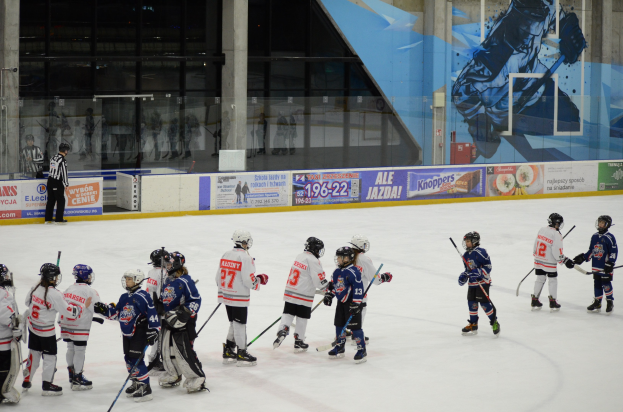 Gruppe junger Menschen in Helmen und Hockey-Ausrüstung, die auf einer Indoor-Eisbahn mit Bannern, Säulen und Glaswänden im Hintergrund Eis hockey spielen.