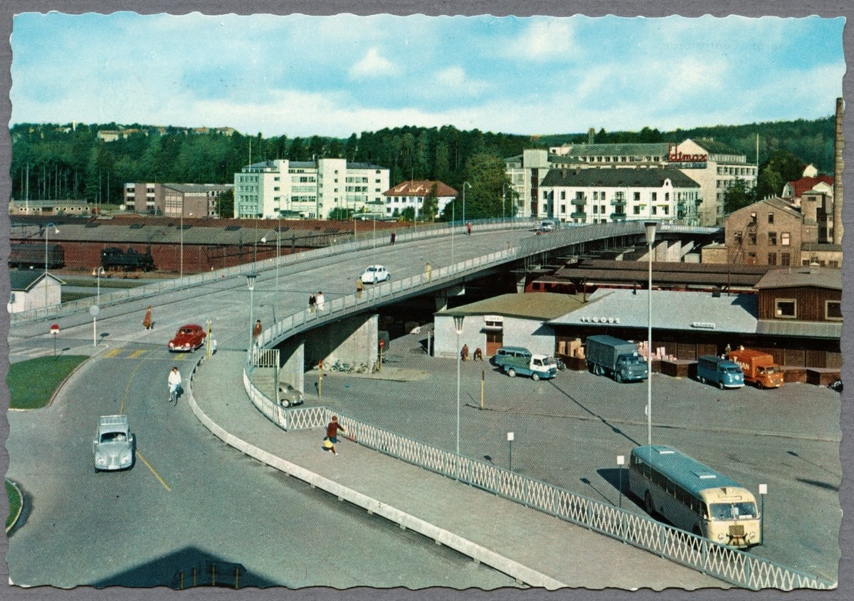 Altes Schwarz-Weiß-Foto einer Stadtstraße mit Autos, Bussen, Fußgängern auf einer Brücke, Laternen, mehrstöckigen Gebäuden, Bäumen und einem bewölkten Himmel.