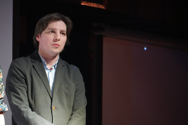 Zwei Menschen, ein Mann im Anzug und eine Frau, stehen Seite an Seite auf einer Bühne mit ernsten Gesichtern.