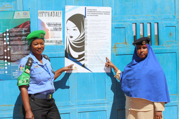 Zwei Frauen stehen nebeneinander vor einer blauen Tür, die mit Plakaten bedeckt ist. Eine trägt ein blaues Hemd, schwarze Hosen und eine grüne Mütze, die andere hält ein Papier in der Hand, was darauf hindeutet, dass sie Teil einer Polizeikraft sind.