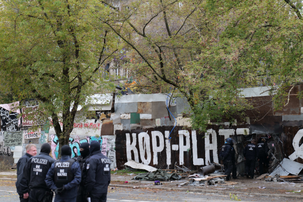 Eine Gruppe von Polizisten in schwarzen Uniformen und Helmen steht vor einem verfallenen Gebäude mit Graffiti, umgeben von verstreuten Trümmern und Bäumen im Hintergrund.