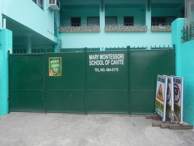 Außenansicht der Mary Montessori School of Cavite, mit einem grünen Tor mit einem Schild, Fenstern, Geländern, Säulen, Klimaanlagen und Informationsschildern auf dem Boden.