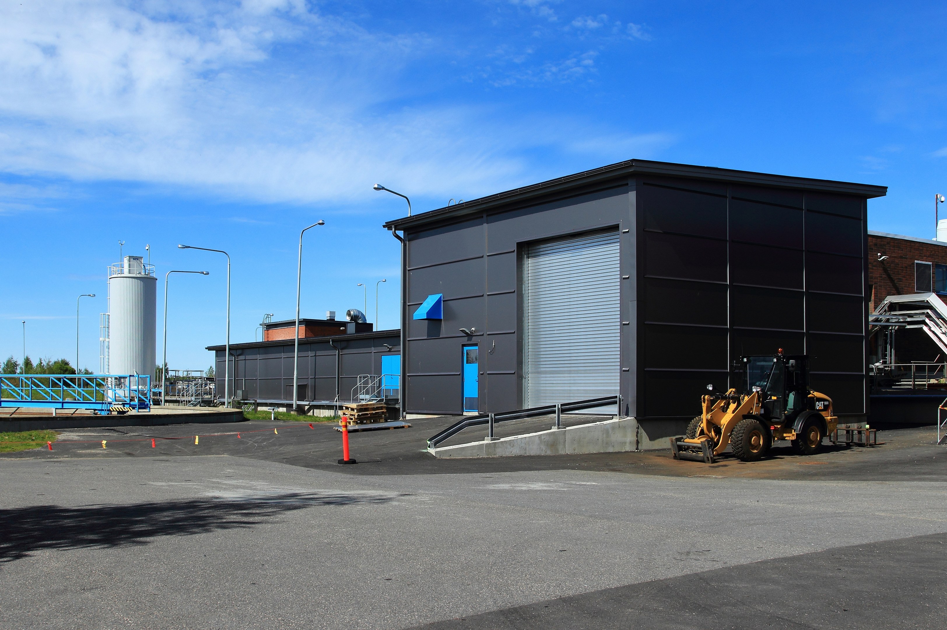 Großes Industriegebäude mit Bulldozer davor, umgeben von Straßeninfrastruktur und Fahrzeugen, unter einem bewölkten Himmel.