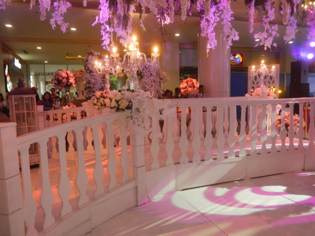 Weiße Reling mit Blumen und Kronleuchtern bei einer Hochzeitsfeier, Menschen umringen die Reling, Säulen und Lichter im Hintergrund, Bildschirm auf der rechten Seite.