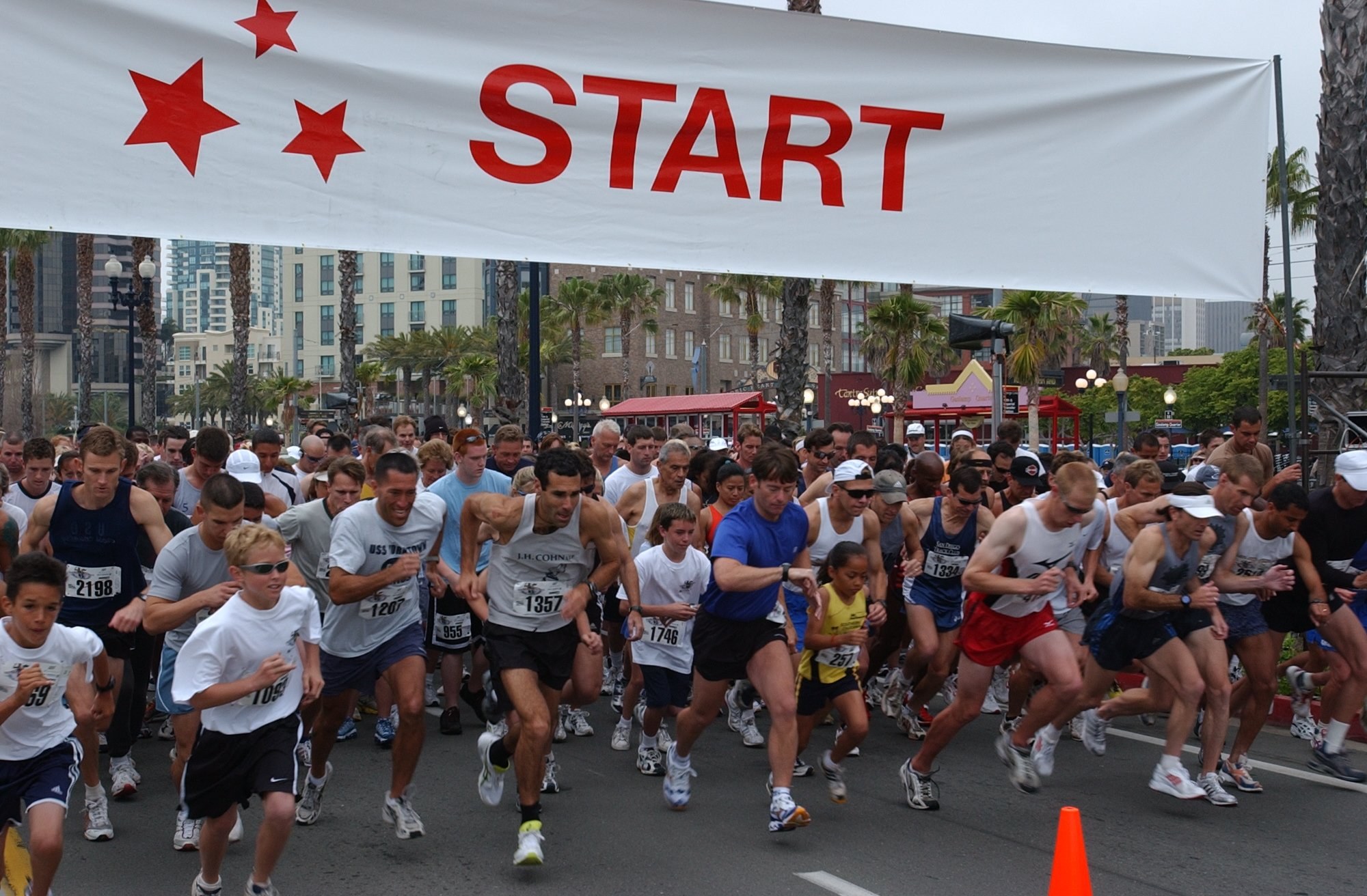 Gruppe von Läufern bei einem Marathon, die an einer Verkehrsinsel vorbeilaufen, mit einem Banner und Gebäuden im Hintergrund bei klarem blauem Himmel.