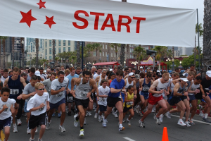 Gruppe von Läufern bei einem Marathon, die an einer Verkehrsinsel vorbeilaufen, mit einem Banner und Gebäuden im Hintergrund bei klarem blauem Himmel.