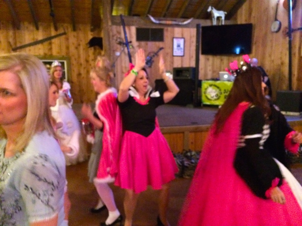 Eine Gruppe von Menschen in Kleidern tanzt in der Mitte eines Raums, während eine andere Gruppe links davon steht, ein Fernseher, ein Spielzeugtier und verschiedene Gegenstände im Hintergrund zu sehen sind.