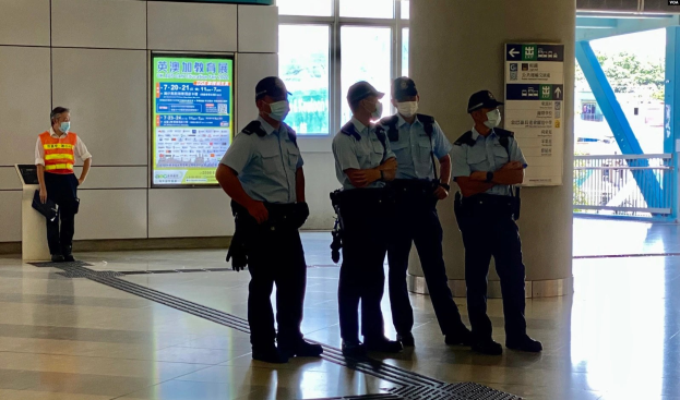 Eine Gruppe von Polizisten in Masken steht vor einem Gebäude mit Glastüren, textbedeckten Tafeln, einer Säule und einem Geländer, mit Bäumen und Fahrzeugen im Hintergrund sichtbar.