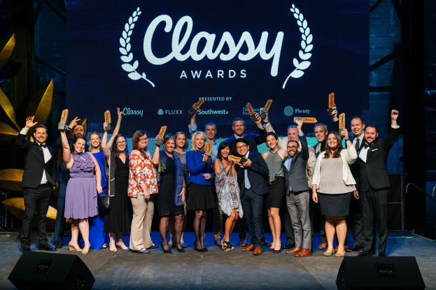 Gruppe von Menschen auf einer Bühne, die Auszeichnungen halten, mit Lautsprechern darunter und einem Bildschirm im Hintergrund, der "Classy Awards 2019" anzeigt.