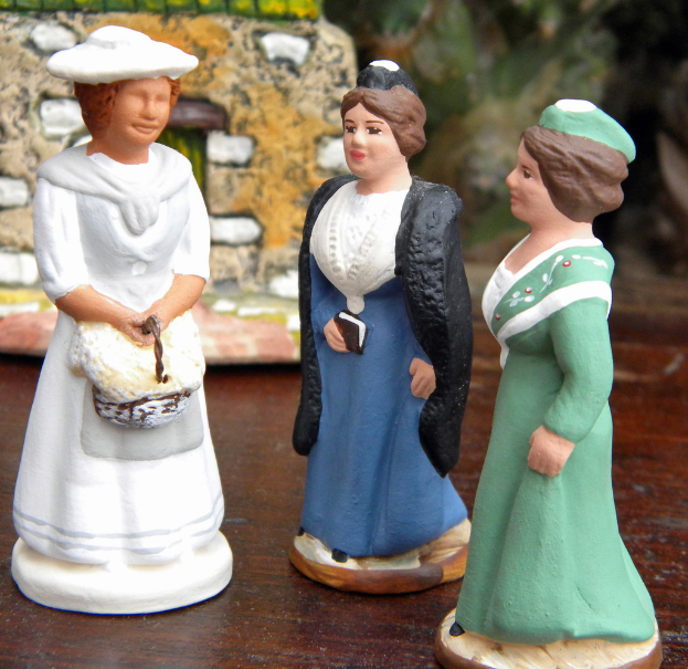 Drei Frauen-Skulptur-Puppen auf einer Holzplatte mit Spielzeughäusern im Hintergrund.