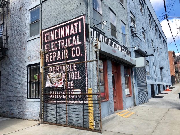 Das Geb├Ąude der Cincinnati Electrical Repair Co. mit Fenstern, T├╝ren, einer Schanktafel, einem Metallzaun, einem M├╝llk├Ârper, Pfosten, Dr├Ąhten, B├Ąumen und einem bew├Âlktem Himmel.