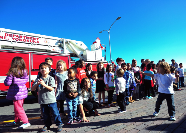Kinder vor einem Feuerwehrauto mit der Aufschrift 'Yokosuka Fire Department's Christmas Parade' auf einer Straße mit Laternenmösten, Bäumen und einem klaren blauen Himmel.