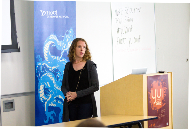 Eine Frau in einem schwarzen Kleid steht neben einem Rednerpult in einem Konferenzraum, mit einem Tisch neben dem Rednerpult und einer Wand, einem Fenster und einem Banner im Hintergrund.