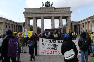 Eine Gruppe von Menschen steht vor dem Brandenburger Tor in Berlin, Deutschland, mit Schildern und Fahnen, auf denen "Fukushima Hiroshima Nagasaki Atomod" gegen die Atombombe protestiert wird, mit Fahrzeugen, Bäumen und einem bewölkten Himmel im Hintergrund.