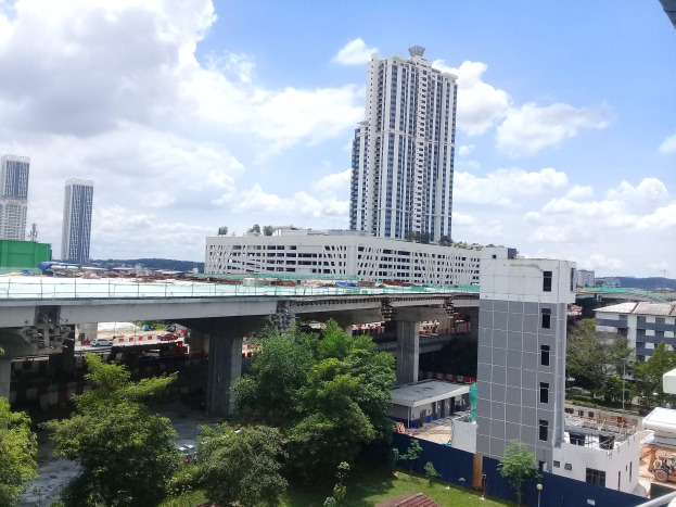 Luftaufnahme einer Stadtlandschaft mit Gebäuden, Bäumen, Gras, Fahrzeugen, einer Brücke, Strommasten und einem bewölktem Himmel, die eine Baustelle für ein neues Wohnkomplex zeigt.