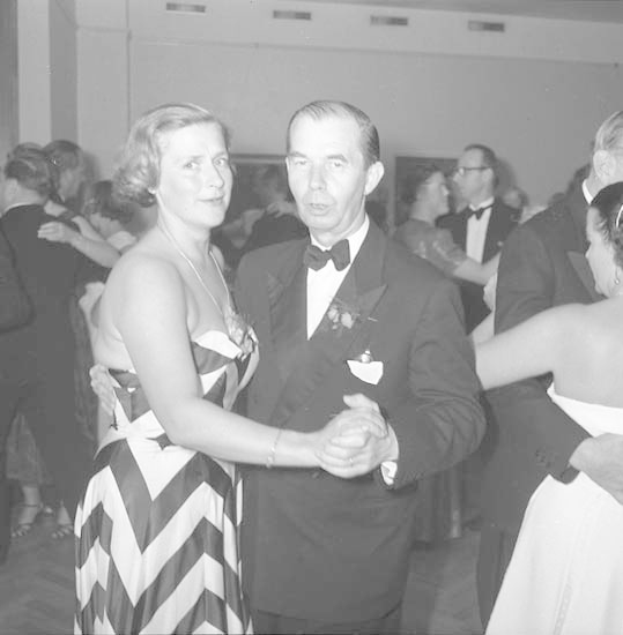 Ein Mann in einem schwarzen Blazer, weißem Hemd und schwarzen Hosen tanzt mit einer Frau in unbekannter Kleidung auf einer formellen Veranstaltung, indem sie sich an den Händen halten und zur Musik schaukeln, vor einer Wand als Hintergrund.