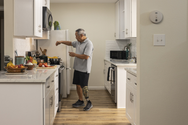 Ein Mann steht in einer Küche mit weißen Schränken und hält ein Glas in der Hand, während verschiedene Küchenutensilien, einschließlich Obst und Gemüse, auf einer Arbeitsplatte zu seiner Linken zu sehen sind.