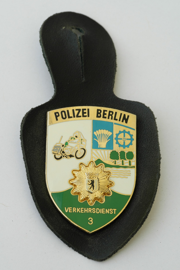 Ein schildförmiges Polizeibadge mit der Aufschrift 'Polizei Berlin' auf einem blauen Hintergrund, einem weißen Rand, einem zentralen weißen Stern und schwarzer fetter Schrift, das auf einer Oberfläche liegt.