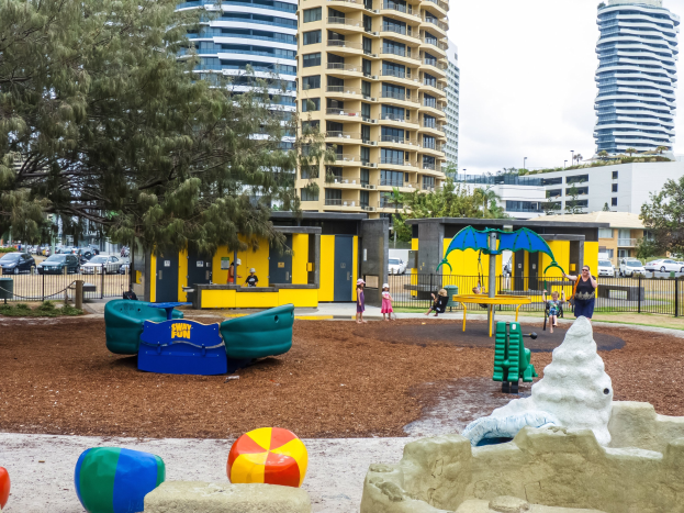 Ein Spielplatz in einer städtischen Umgebung mit spielenden Kindern auf der Ausrüstung, umgeben von Bäumen und hohen Gebäuden im Hintergrund.