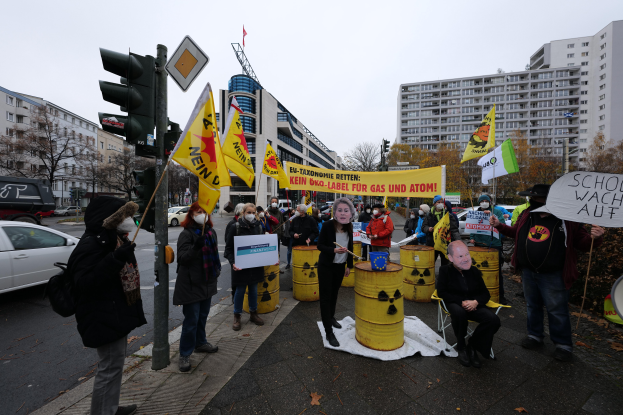 Eine Gruppe von Menschen steht auf einer Straße und hält gelbe Fässer und Schilder hoch, mit einer Person im Vordergrund, Fahrzeugen, Verkehrssignalen, Hinweistafeln, Bäumen, Gebäuden und einem klaren blauen Himmel im Hintergrund, die gegen Atommüll protestieren.
