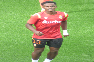 Emmanuel Adebayor, ein Mann in rotem T-Shirt und schwarzen Shorts, geht auf einem Fussballfeld mit zwei Personen im Hintergrund.