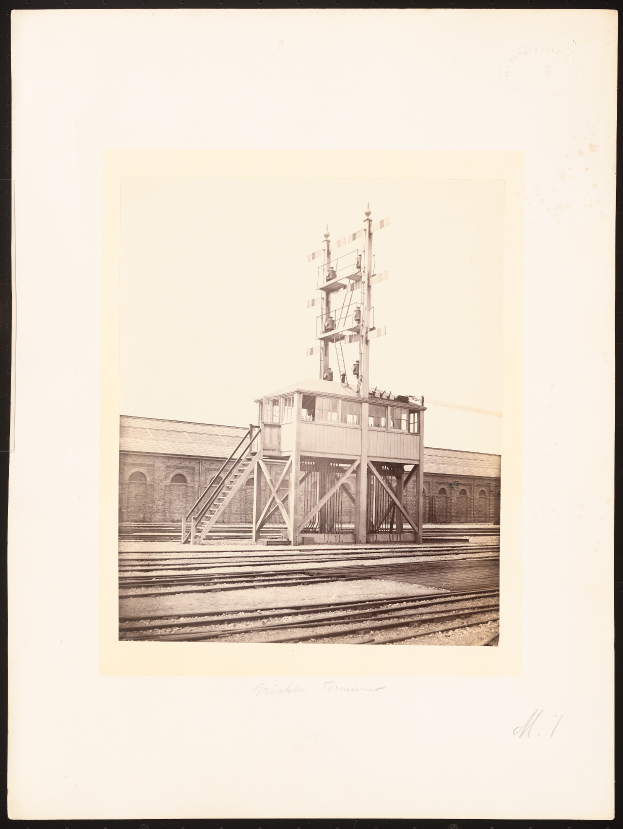 Ein Schwarz-Weiß-Foto eines Bahnhofs mit einer zentralen Signalbox, umgeben von Bahnschienen und einem Gebäude im Hintergrund, mit Text unten.