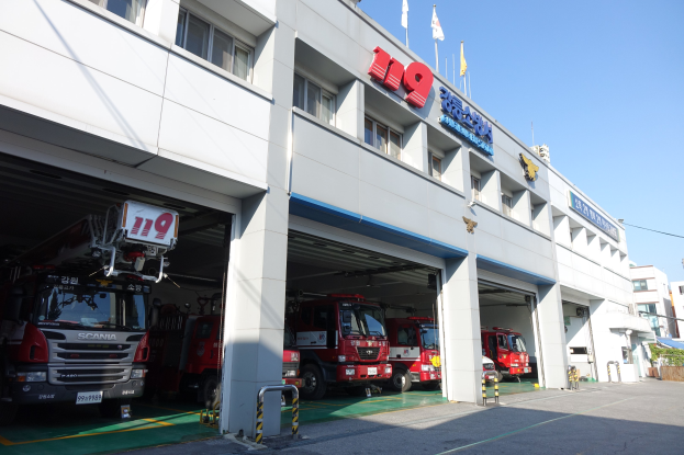 Feuerwache mit mehreren Feuerwehrautos vor der Tür, mit Fenstern, Säulen, Namensschildern und Flaggen im Vordergrund; andere Gebäude sind im Hintergrund unter einem klaren blauen Himmel zu sehen.