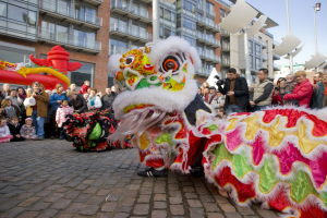 Eine Löwen-Tanzvorstellung während einer Chinesischen Neujahrsfeier in Amsterdam, mit einer Menge Menschen, die zusehen und den Moment mit Kameras festhalten, vor einer Kulisse aus Gebäuden und einem klaren blauen Himmel.