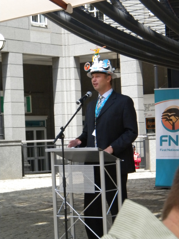 Ein Mann im Anzug und Hut spricht in ein Mikrofon an einem Rednerpult, mit einer Person rechts unten, einem Banner mit Text rechts und einem Gebäude mit Geländer im Hintergrund.