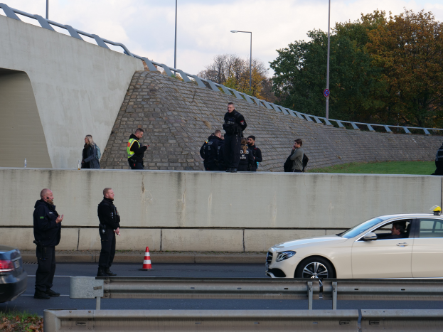 Eine Gruppe von Polizeibeamten steht neben einem Auto auf der Straße, mit Verkehrskegeln, einer Begrenzung, Gras, einer Wand, Laternenpfählen, Bäumen und einem bewölkten Himmel im Hintergrund.