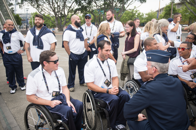 Eine Gruppe von Menschen in Rollstühlen, die vor einer Kulisse aus Bäumen, Pfählen, Gebäuden und einem klaren blauen Himmel miteinander sprechen, wobei einige Mützen und Brillen tragen.